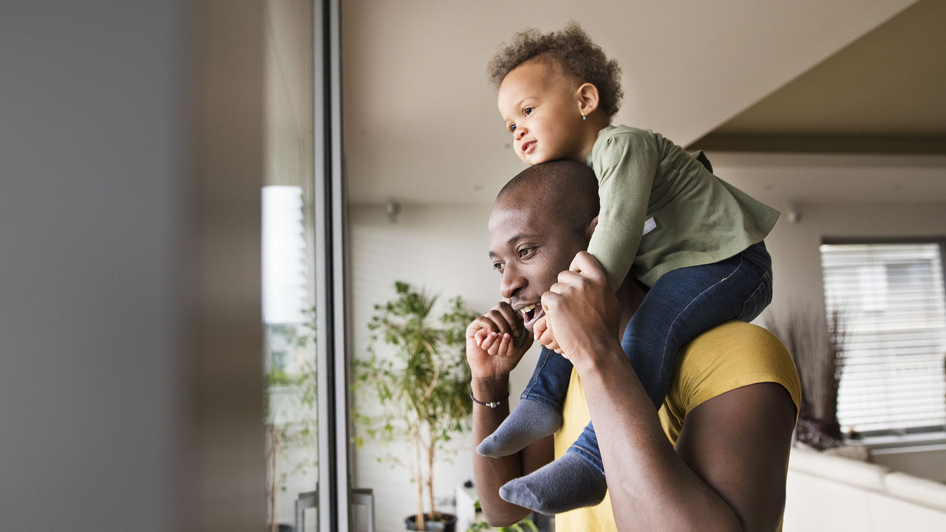 Man carrying a young child on his shoulders looks out the window of his home